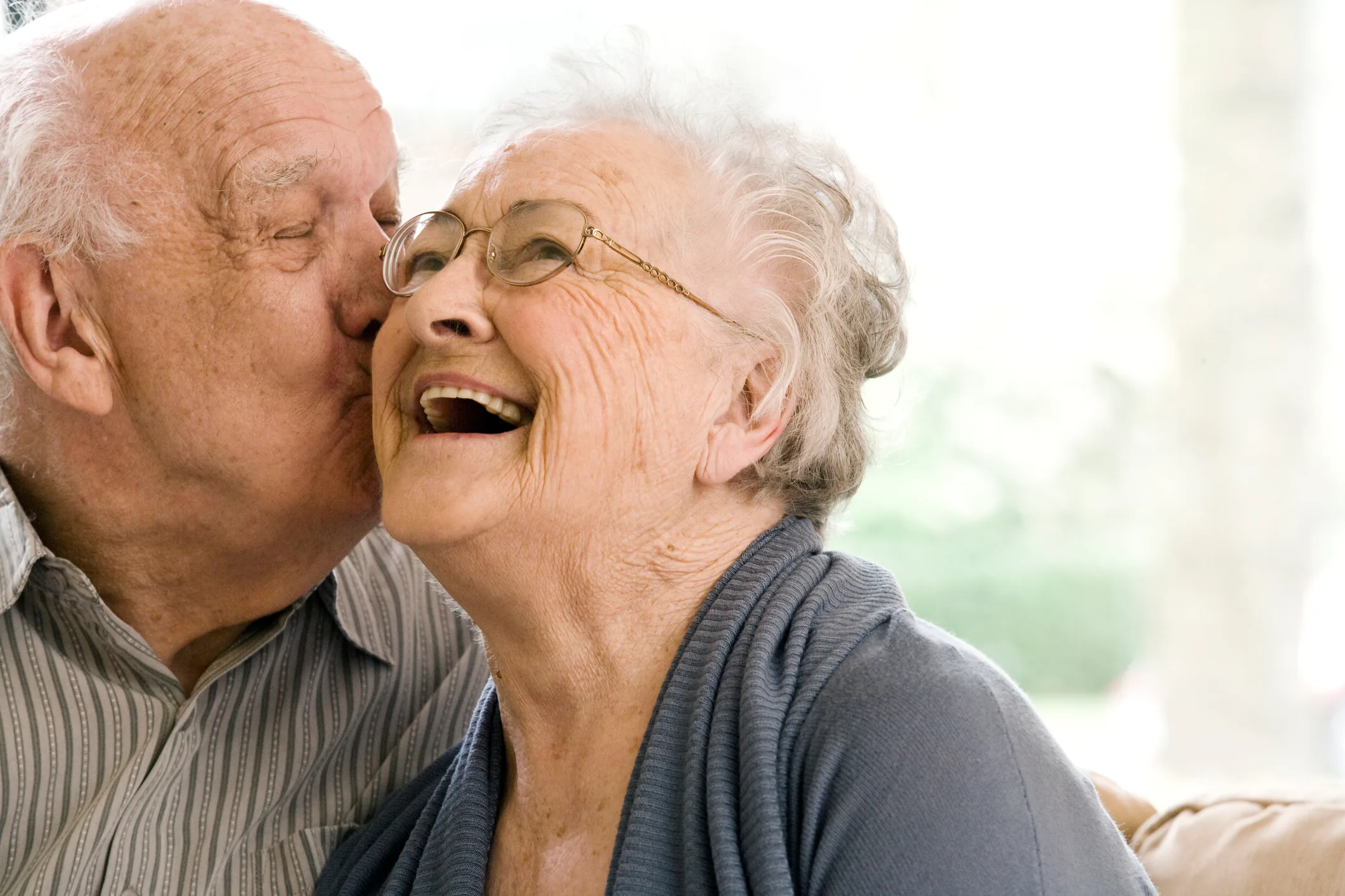 Senior couple at home. The husband is kissing his wife's cheek while she laughs with her eyes open.